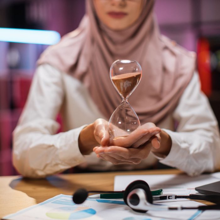 Focus on hands of businesswoman sitting at desk with modern laptop and holding hourglass